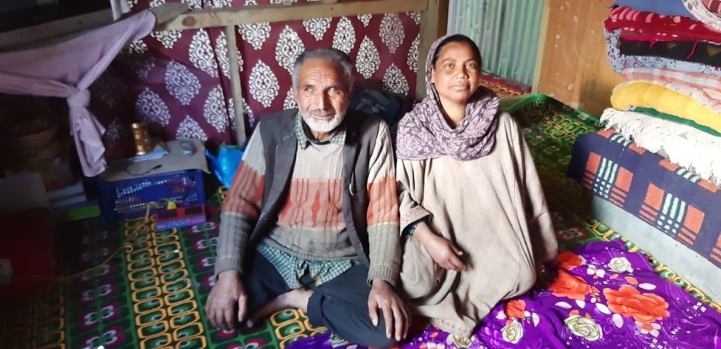 An elderly couple sitting on a colorful patterned floor in a cozy indoor setting, with traditional decor around them.