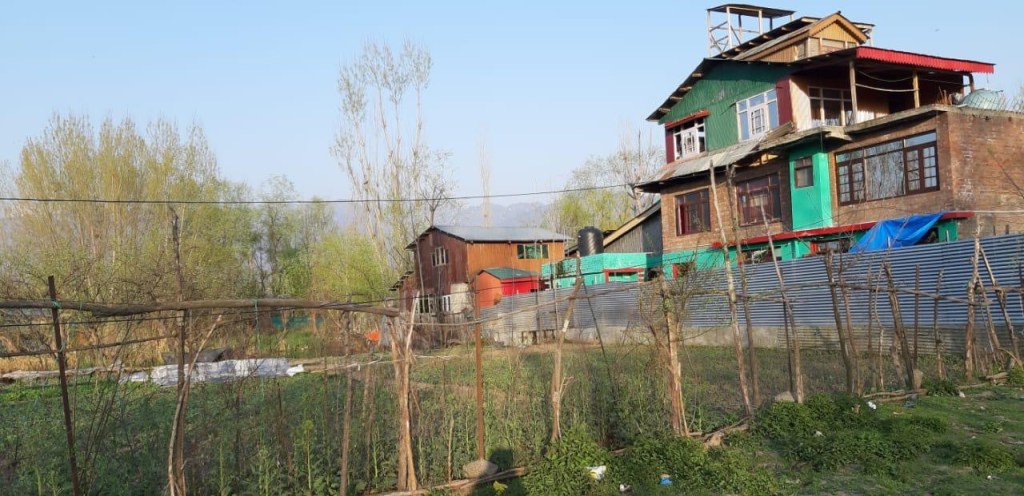 A scenic view of a rural area featuring various buildings, including a multi-story house with green and red accents, surrounded by trees and agricultural land with a fence.