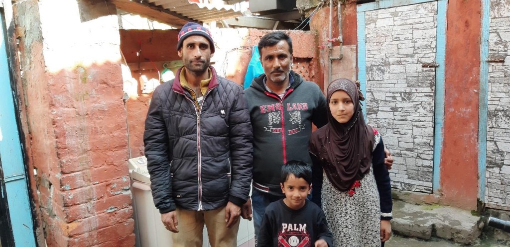 A family of four poses together in a narrow outdoor space, with a backdrop of weathered walls and doors. Two adult men and two children, a girl and a boy, are dressed in casual clothing.
