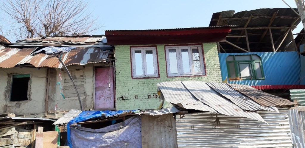 An image of a multi-level structure with a mix of corrugated metal roofing and brick walls, featuring a green house with white windows and a pink door, surrounded by makeshift materials.