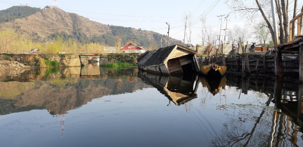 A partially submerged wooden house leaning in a river with reflections visible on the water's surface, surrounded by hills and trees.