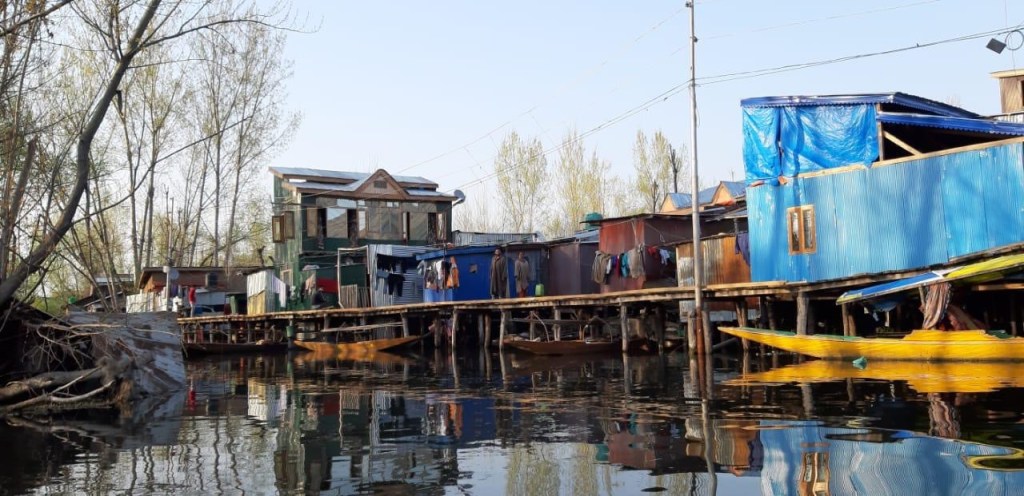 Colorful houseboats along a waterway with reflections in the calm water, surrounded by trees.